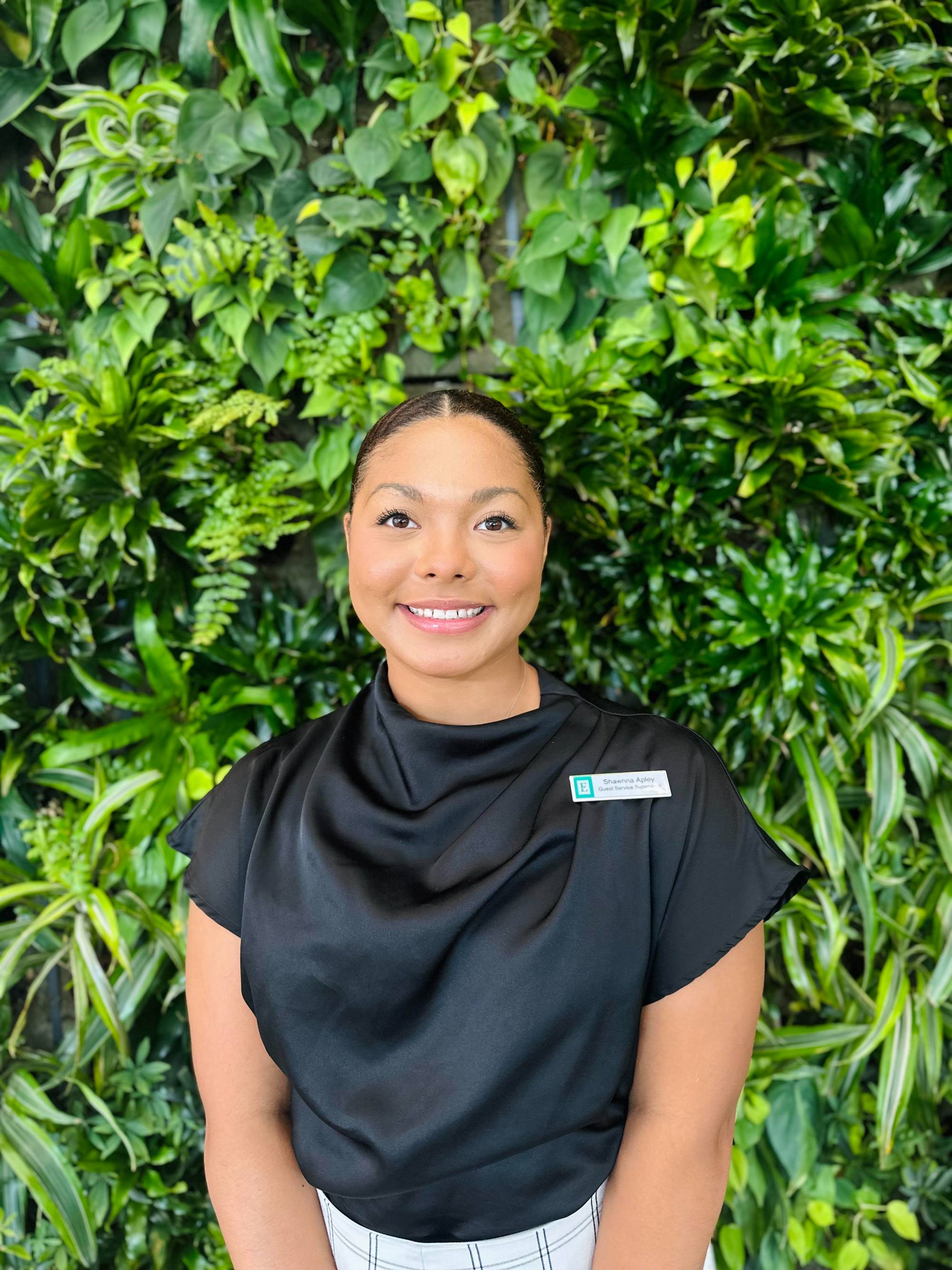 Photo of African American woman in a black shirt standing in front of a wall of plants